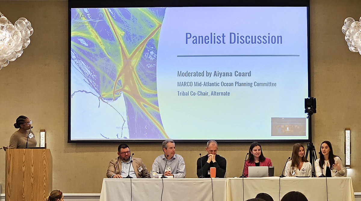 A panel of speakers seated at a table with a slide behind them reading "Panelist Discussion"